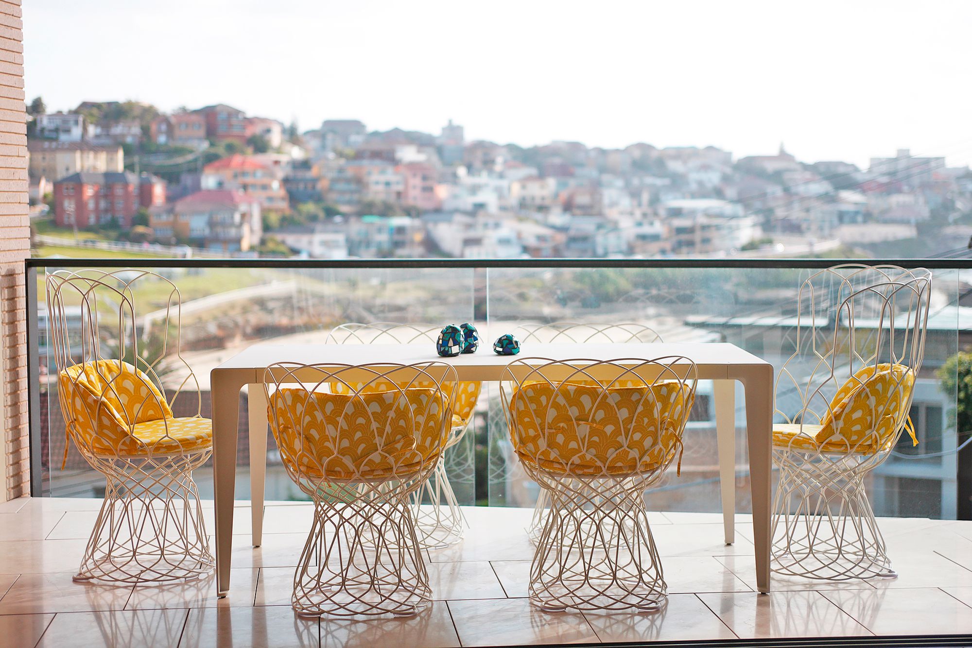 balcony dining area with wire chairs and yellow cushions overlooking coastal village and ocean
