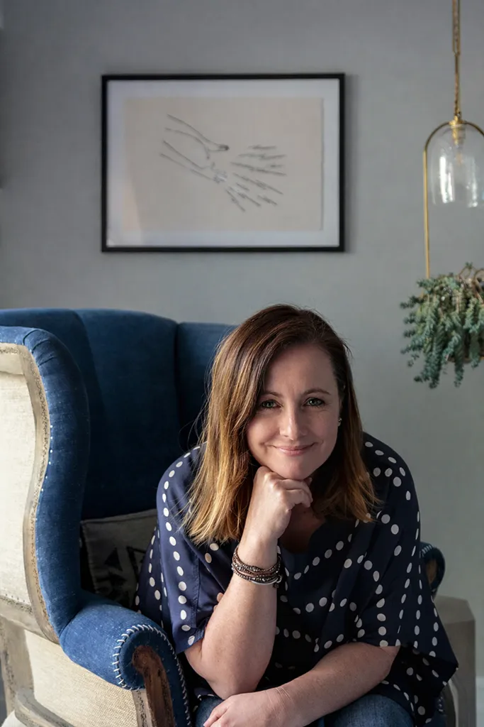 Bronwyn Poole, Los Angeles interior designer, seated in blue wingback chair with abstract art and brass pendant light