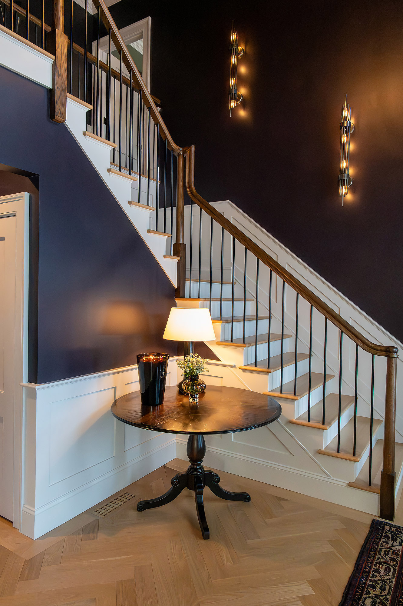 Foyer staircase aubergine walls wainscot vertical sconces pedestal table lamp herringbone floor — Bronwyn Poole Interiors