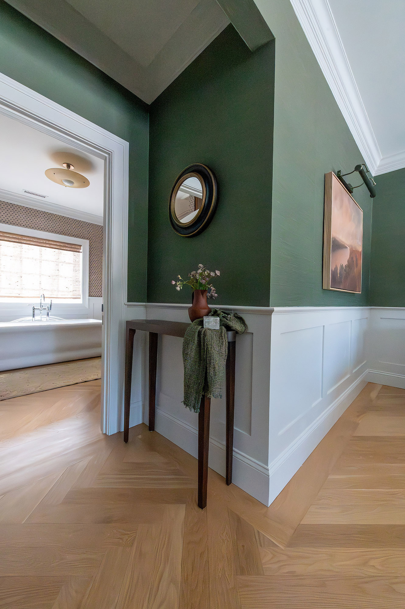 Primary bedroom hallway green walls white wainscot walnut console convex mirror landscape painting herringbone floor — Bronwyn Poole Interiors