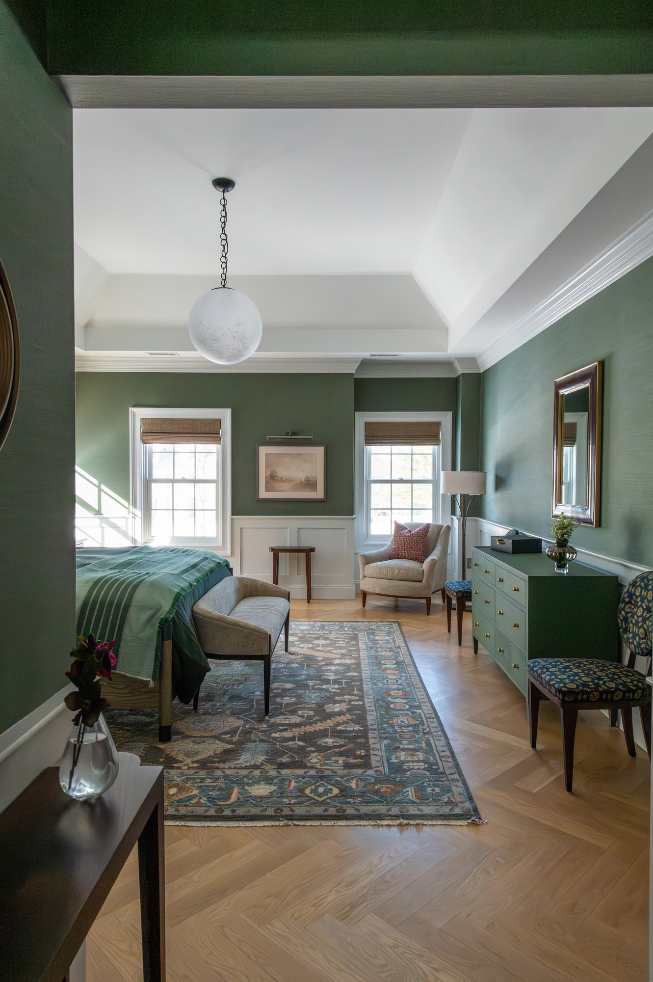 Primary bedroom wide view tray ceiling globe pendant green grasscloth walls white wainscot river landscape art velvet armchair block print side chair persian rug herringbone floor — Bronwyn Poole Interiors