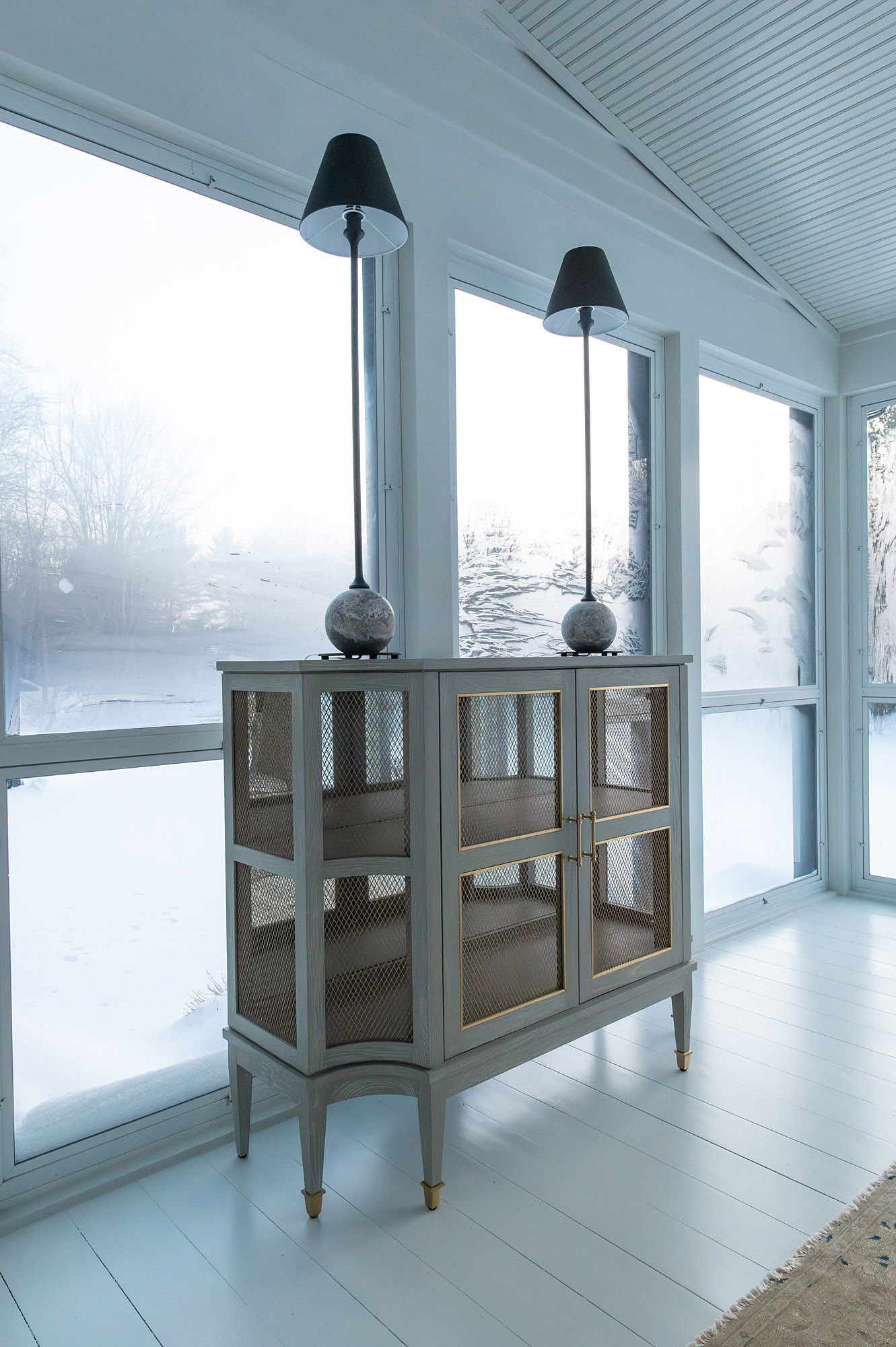 Sunroom cane cabinet pair stick lamps stone orb bases brass hardware snowy window view white floor — Bronwyn Poole Interiors
