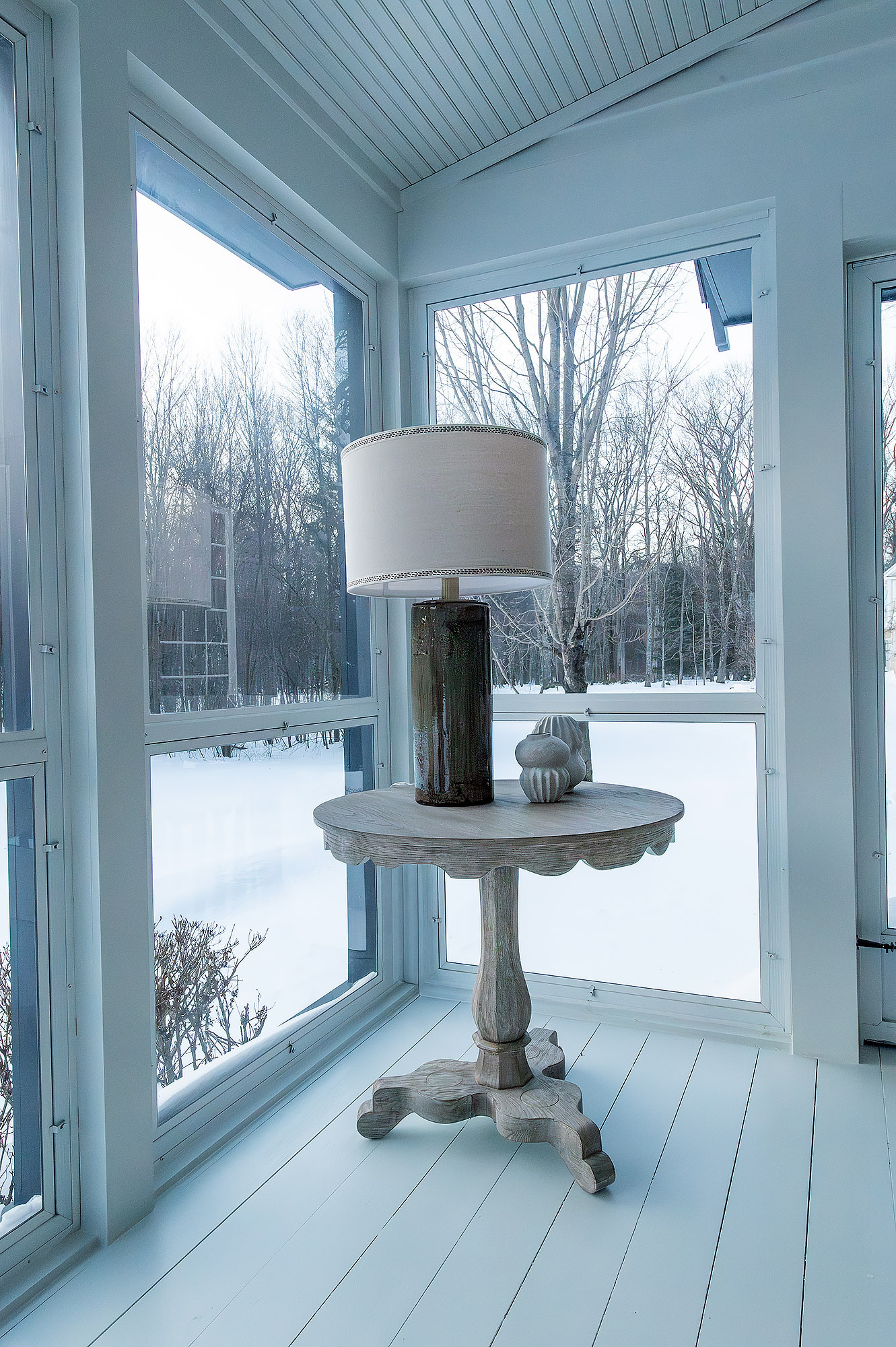 Sunroom corner detail scalloped edge pedestal table bronze cylinder lamp linen drum shade fluted stone finial snowy window view — Bronwyn Poole Interiors