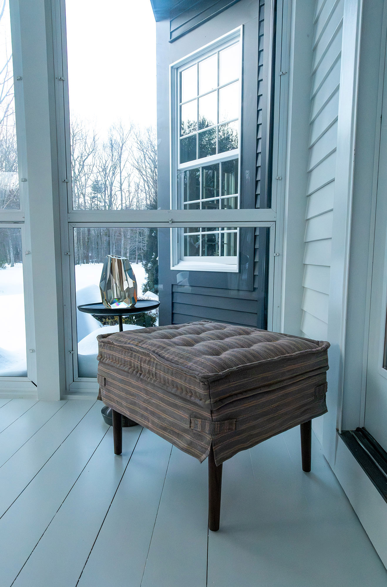 Sunroom corner vignette striped tufted ottoman faceted glass lamp black side table snowy window view — Bronwyn Poole Interiors
