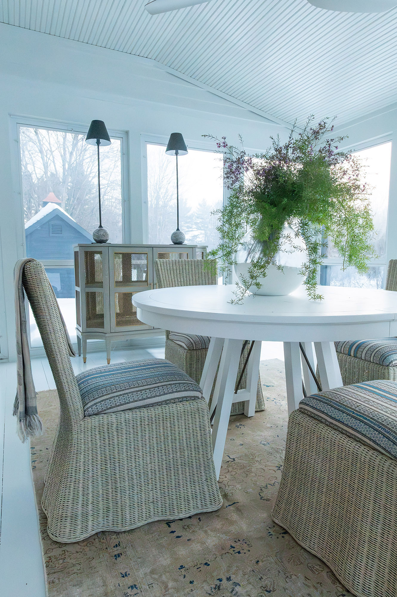 Sunroom dining area white pedestal table skirted wicker dining chairs cane cabinet stick lamps asparagus fern snowy window view — Bronwyn Poole Interiors