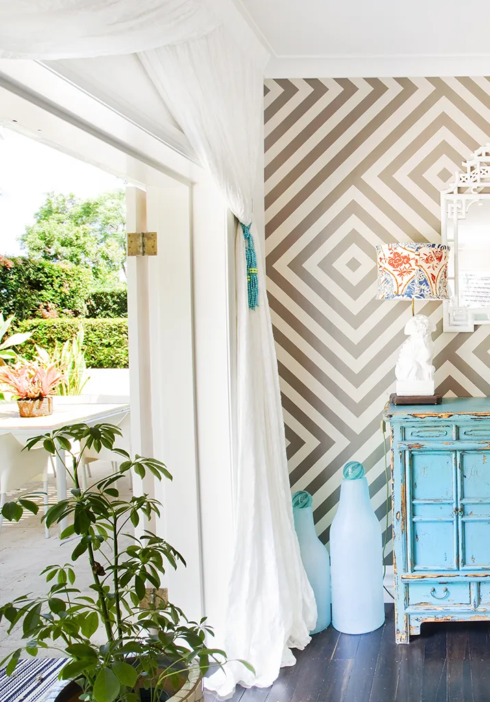 Geometric chevron wallpaper with flowing white curtains and turquoise vintage cabinet framing garden view — architectural curtain design by Bronwyn Poole